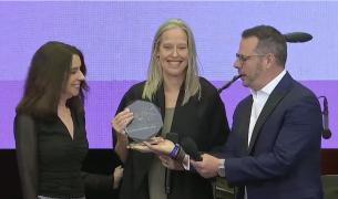 A white woman with long blond hair receives an award from a white man in a suit and a white woman with brown hair 