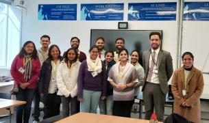 A group of brown-skinned young men and women pose together in front of a classroom