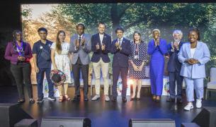 A row of 10 adults of diverse ethnicities stands on a stage clapping in front of an image of leafy treetops