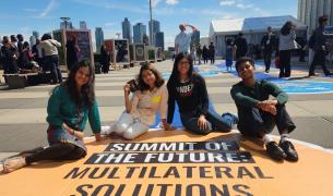 Four diverse young people sit on a yellow street mural that says Summit of the Future Multilateral Solutions with a blue sky and NYC skyline behind them