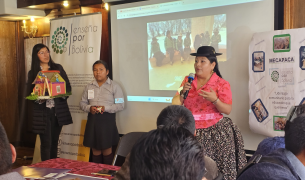 A Bolivian woman in traditional dress wearing a black rimmed hat does a presentation in front of a screen and a poster than says Mecapaca and Ensena por Bolivia