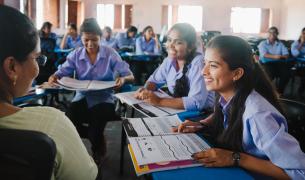 Three you girls sit in desks facing a teacher in a school, they are all smiling