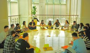 A diverse group of young people and young adults sit in a circle in front of a large window