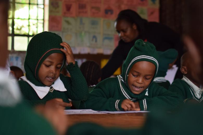 Two Black children in green uniforms with hoods write at a desk as a teacher leans over a desk behind them