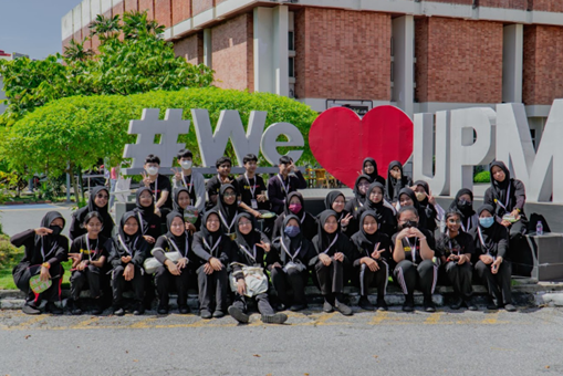 A large group of Asian teenagers, all wearing black, pose in front of a sign that says #We(heart)UPM