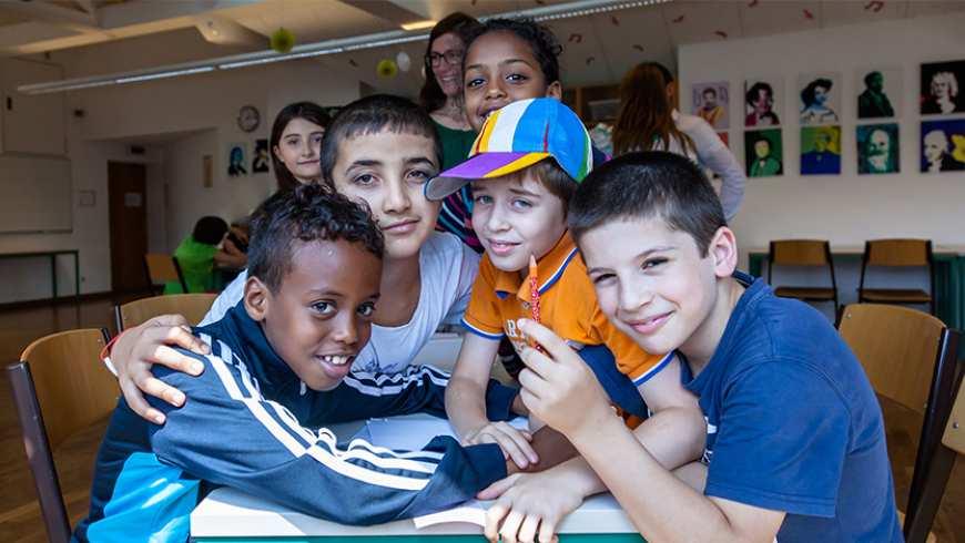 A group of boys of diverse ethnicities sitting at a table with notebooks crowd into the frame with their arms around each other smiling at the camera