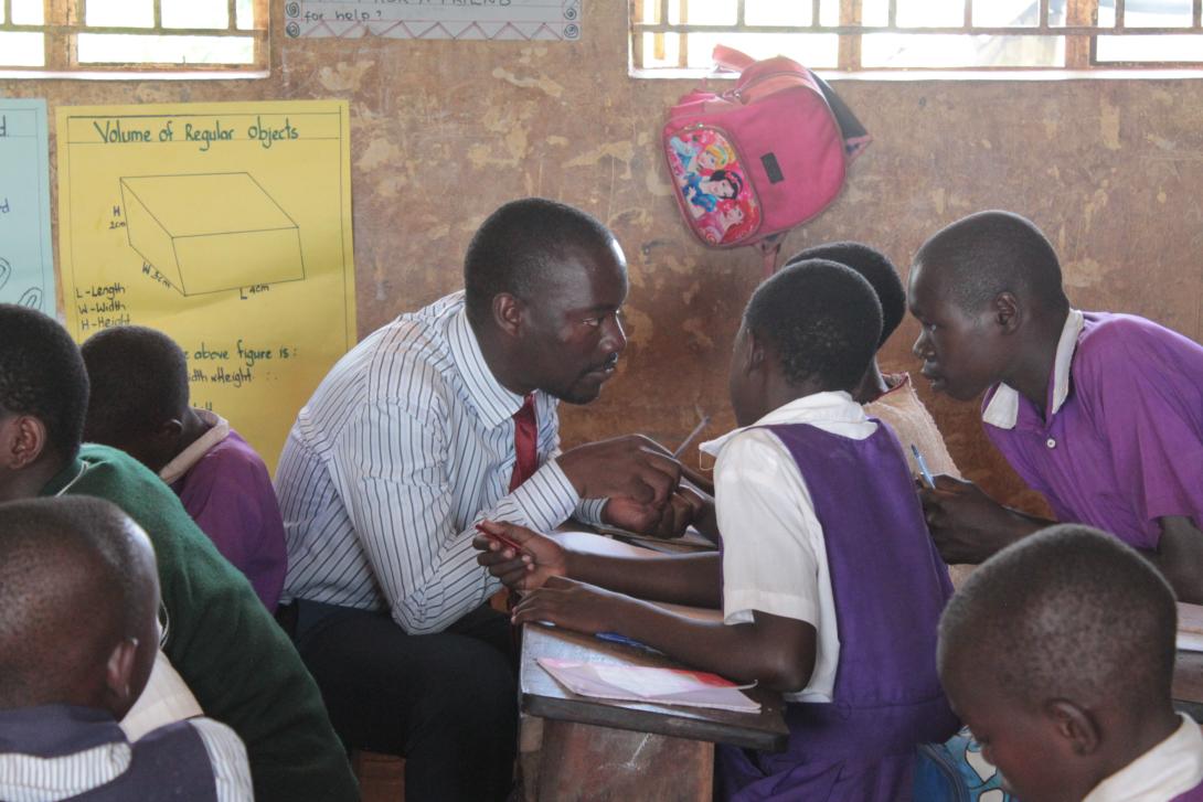 In a clasroom in Uganda, a male teacher wearing a shit and tie speaks with engaged-looking students at their desk