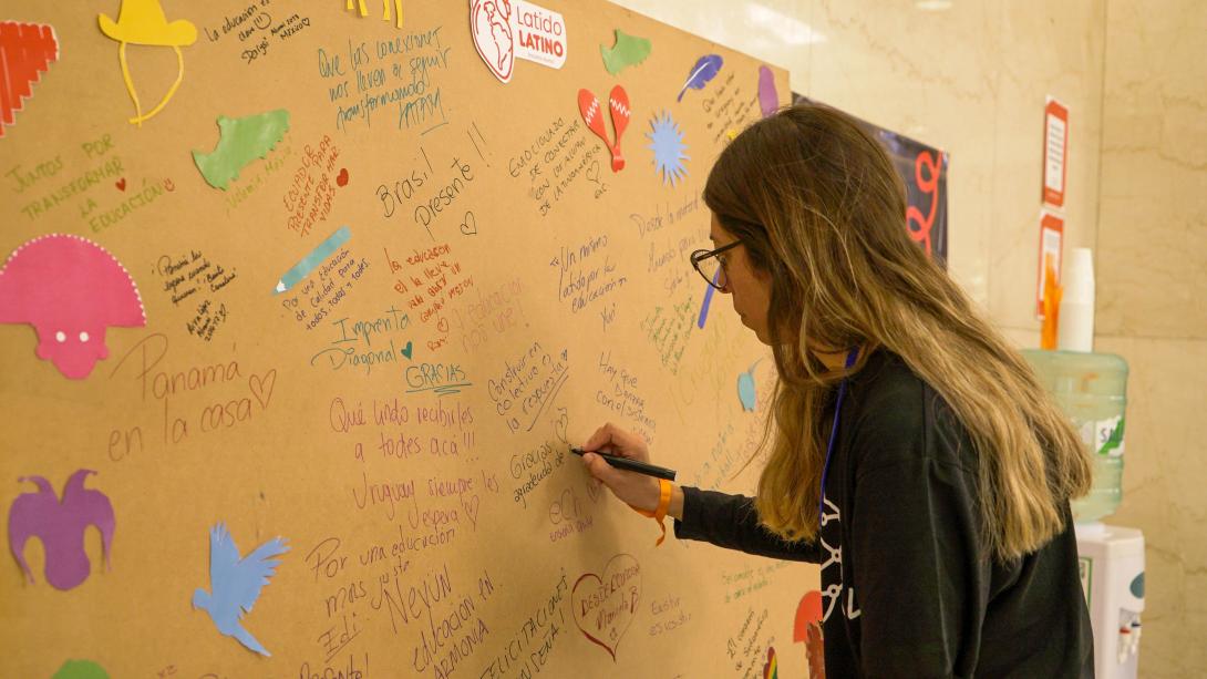 A young woman with long hair writes on a huge piece of paper on a wall with notes from Teach For All Latin American partners