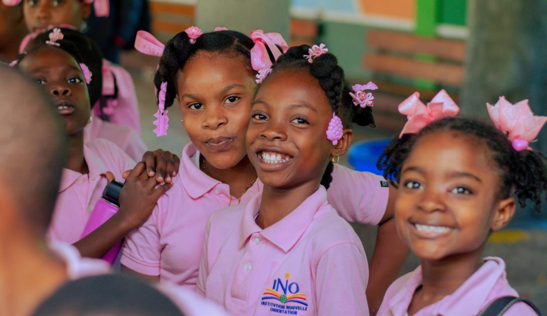 Three Black girls in pink polo shirts with pink bows in their hair smile at the camera