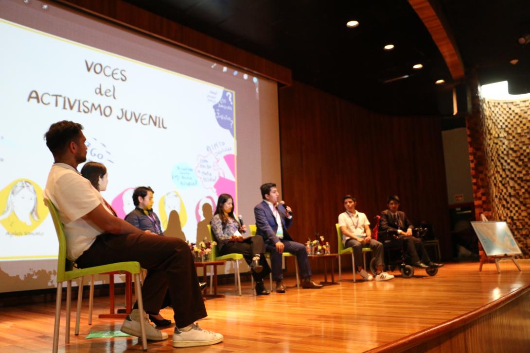 Seven young adults sit in chairs on a stage in front of a screen that says Voces del Acitvismo Juvenil