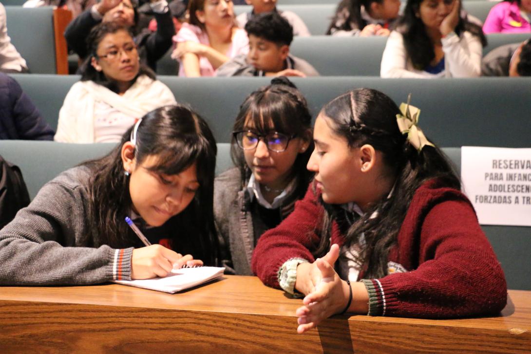 Three Mexican girls in grey and maroon uniforms write on a pad of paper in an auditorium