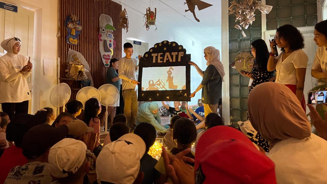 Two young people hold up a shadow puppet theater in front of a room full of others watching, weveral wearing head scarves and caps.