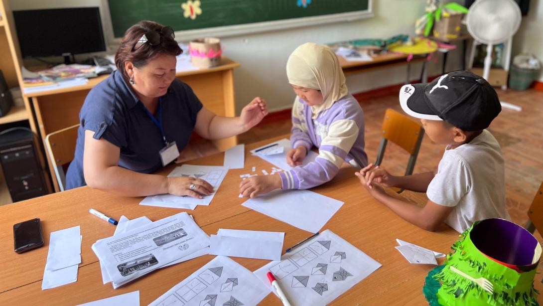 A young female teacher in a blue top shows worksheets to two young students at a table, one wearing a headscarf and the other a baseball cap