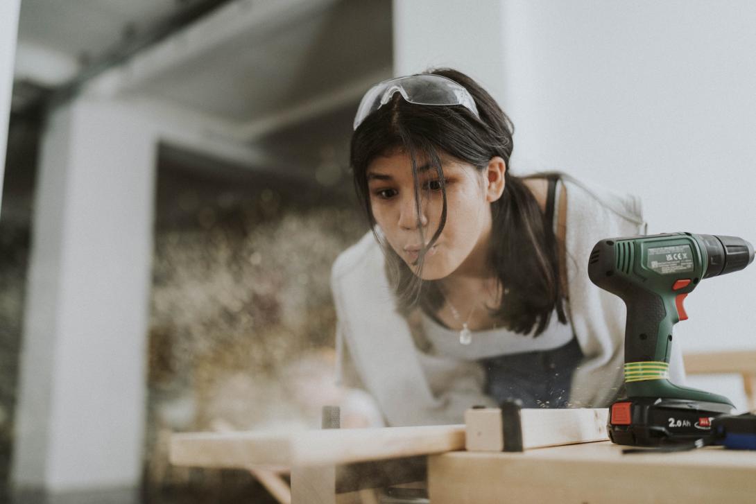 A young teenage girl with brown hair and goggles on her head blows sawdust from a plant next to a powerdrill