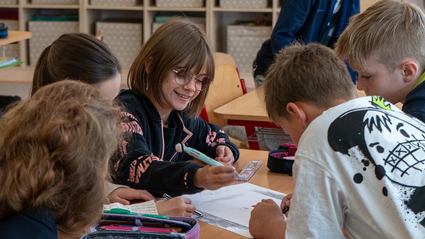 Students cluster around a desk looking and writing in a book