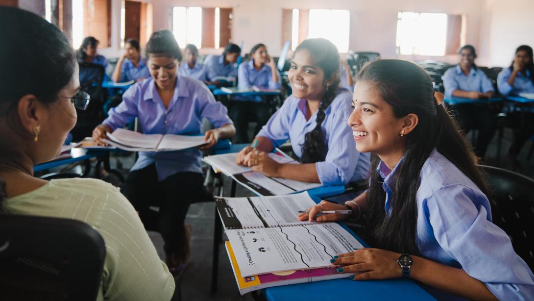 Three you girls sit in desks facing a teacher in a school, they are all smiling